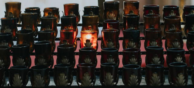 A single devotional candle is lit in a rack of candles in a chapel.